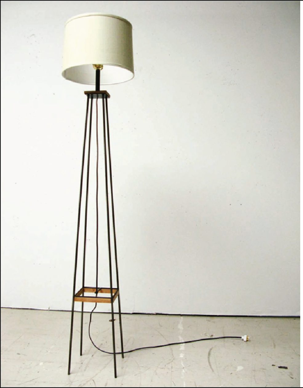 A BILT floor lamp with a slender blackened steel frame, wooden collar and pale drum shade, standing against a bare studio wall.